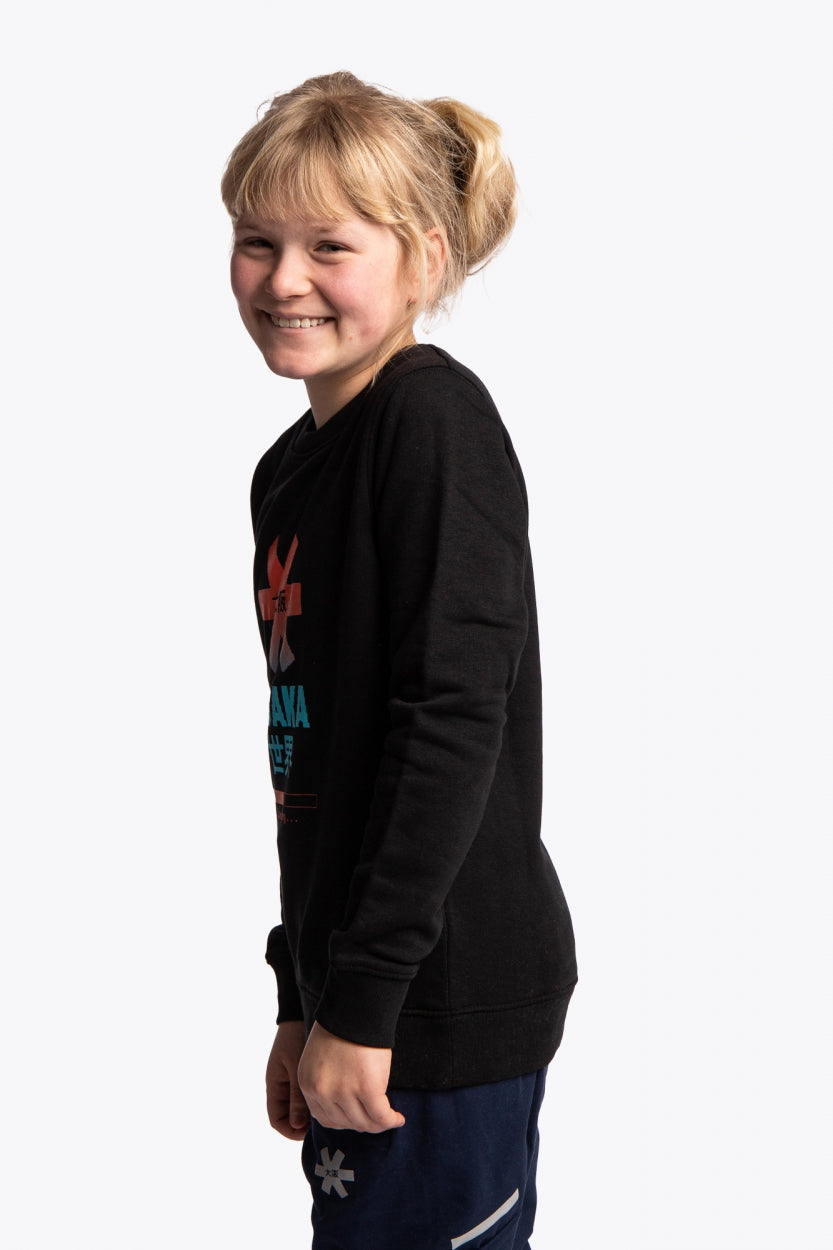 Girl wearing the Osaka kids pixo sweater in black with orange and blue logo. Side view