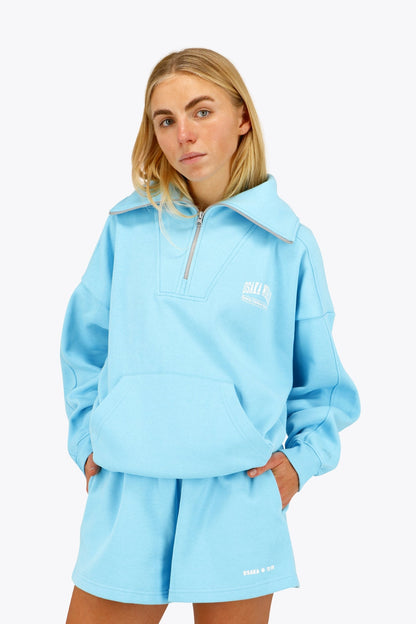 Woman wearing the Osaka women half zip sweater in light blue with white logo. Front view