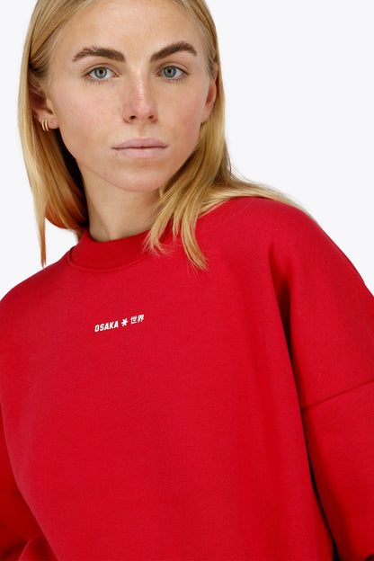 Woman wearing the Osaka women sweater in red with logo in white. Front detail logo view