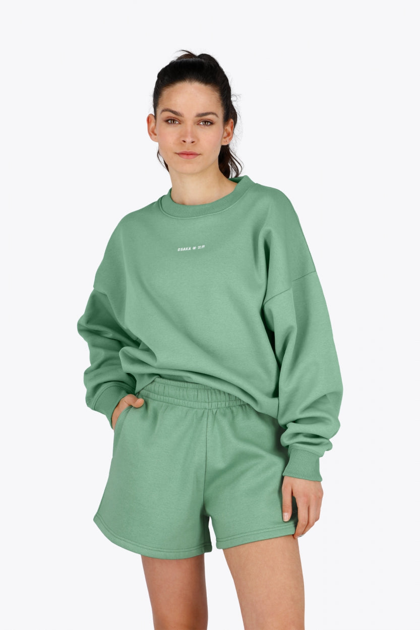 Woman wearing the Osaka women sweater in green with logo in white. Front view