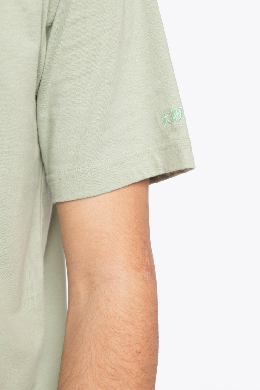 Man wearing the Osaka unisex tee in iceberg green with white logo. Detail sleeve view