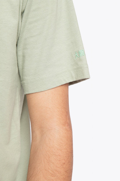 Man wearing the Osaka unisex tee in iceberg green with white logo. Detail sleeve view