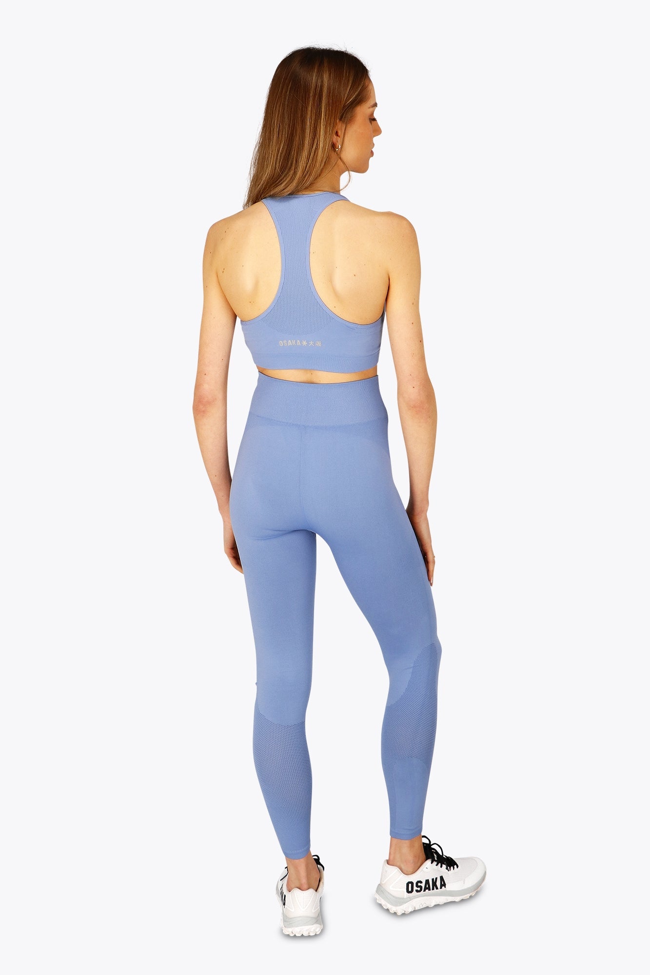 Woman wearing the Osaka women tech sports bra in manor blue with logo in grey. Back view